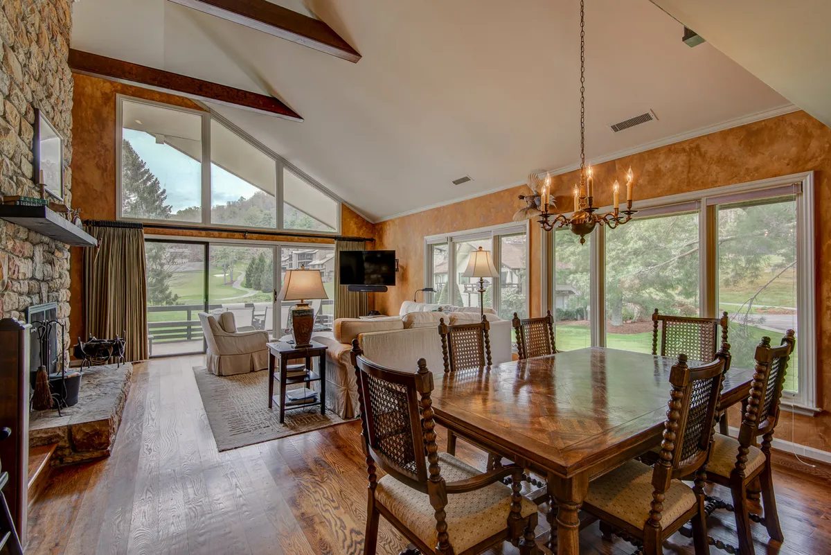 This interior image captures a spacious living area featuring a striking stone fireplace, large south-facing windows, a comfortable seating area, and a dining table. Natural light pours into the room, highlighting the open-concept layout and drawing the eye toward the panoramic views outside.