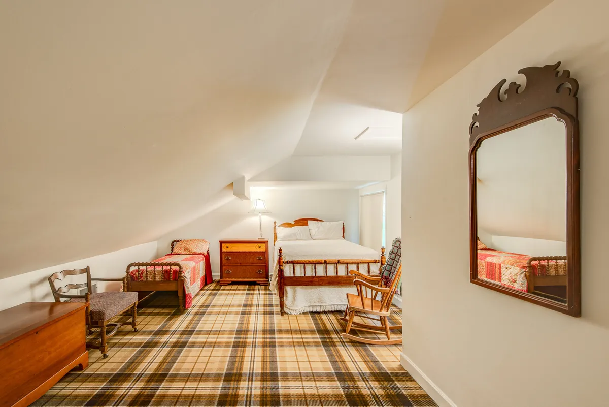 A flexible bedroom setup with two beds, including a full-size and a smaller child-sized bed, a classic wooden rocking chair, and a dressing table with mirror. The plaid carpet and warm lighting create a family-friendly, welcoming atmosphere.