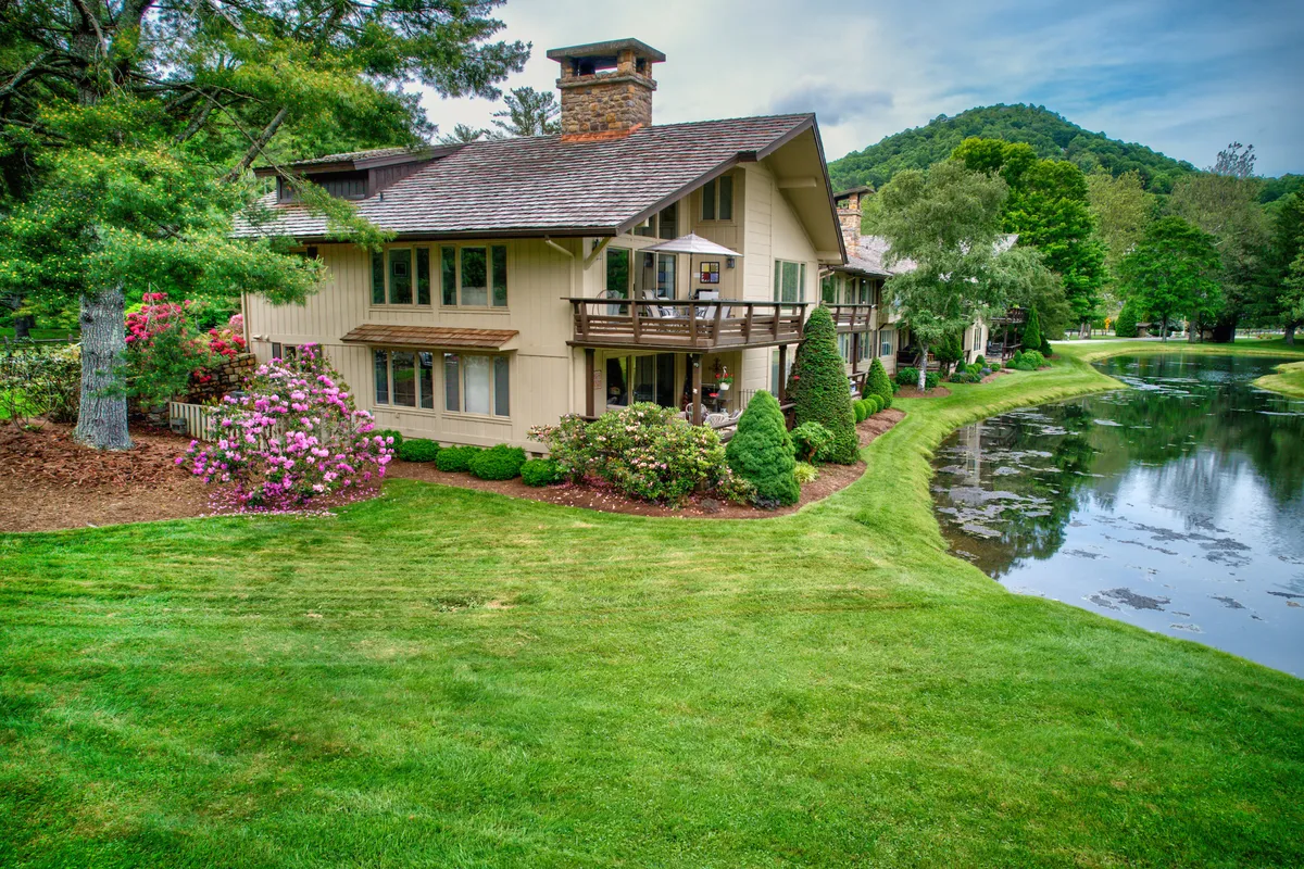 The hero image showcases the exterior of a spacious condo set against the serene backdrop of Boone's lush mountains. The tranquil pond glistens in the foreground, surrounded by a well-kept garden and mature trees, inviting guests to relax, fish, or stroll through nature. The entire scene radiates peace and natural beauty, perfect for unwinding and enjoying time outdoors.