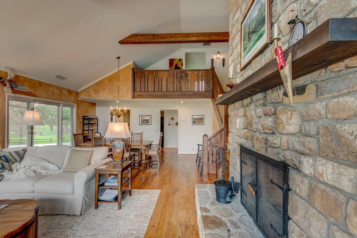 A welcoming, comfortable living and dining space with a stone fireplace, wooden beams, a large window, and a rustic dining table. The atmosphere is cozy and warm, ideal for gatherings and winter evenings.