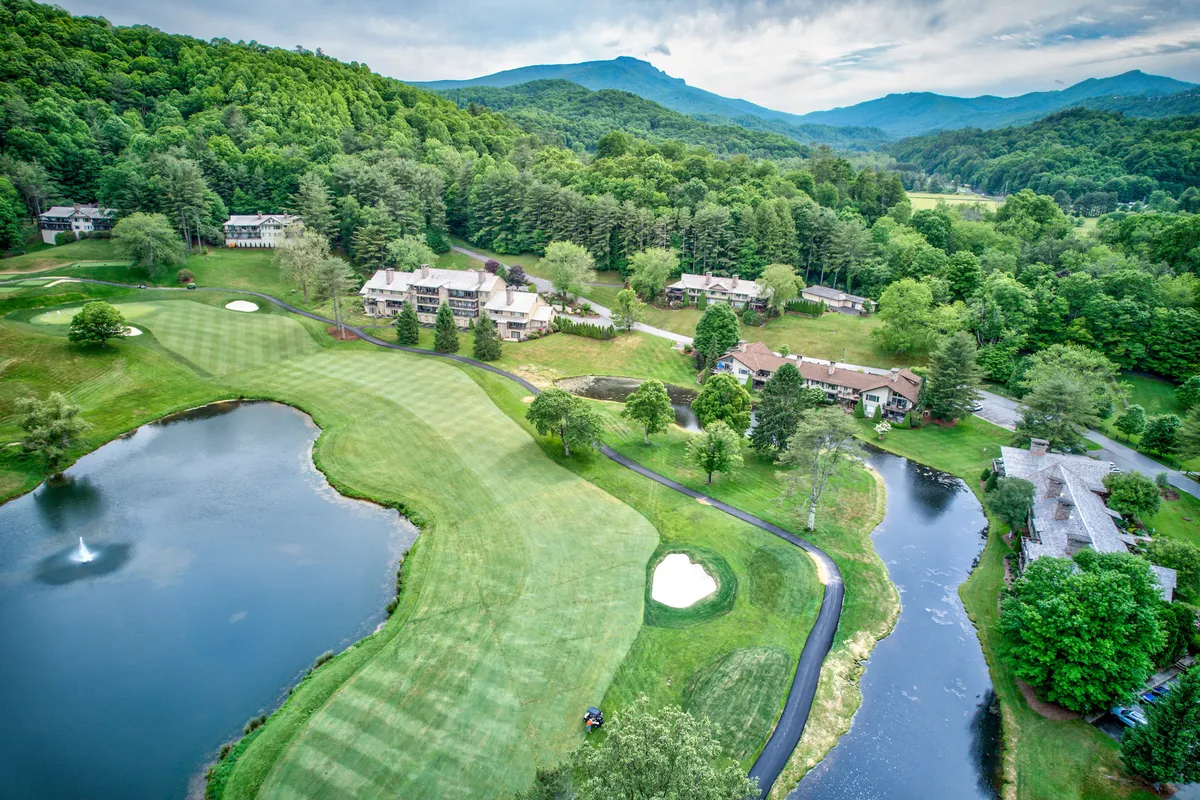 A sweeping exterior view of a lush green golf course bordered by a sparkling pond, with majestic mountains and residential buildings in the background. The serene landscape is enhanced by soft sunset lighting and hints of wildlife near the water’s edge.