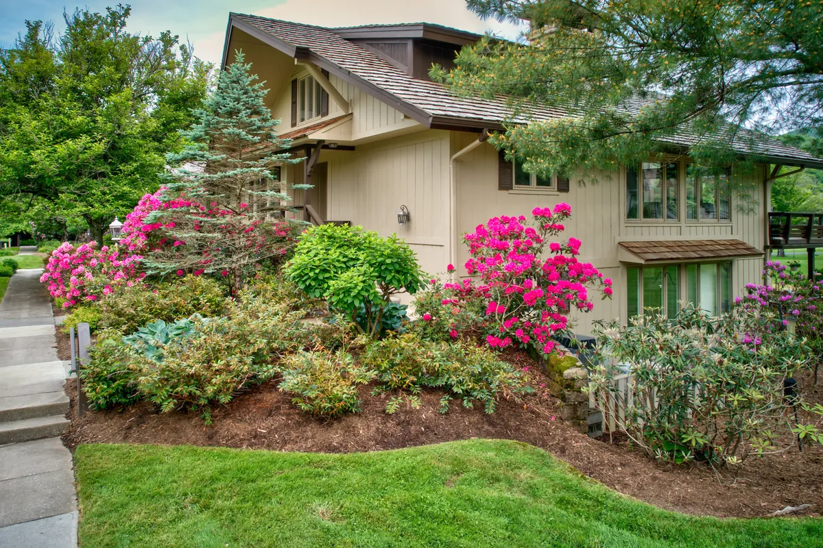 A cheerful garden scene, featuring bright pink flowers, lush landscaping, and a picturesque house nestled in greenery. The outdoor table and seating indicate opportunities for private, romantic meals or quiet relaxation amidst nature.