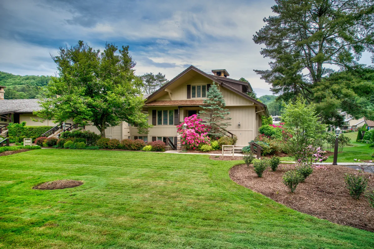 This image features the house enveloped by a lush green lawn, vibrant flower beds, and tall surrounding trees. The welcoming façade, colorful landscaping, and spacious outdoor areas evoke a sense of serenity and space, perfect for group gatherings and outdoor activities.