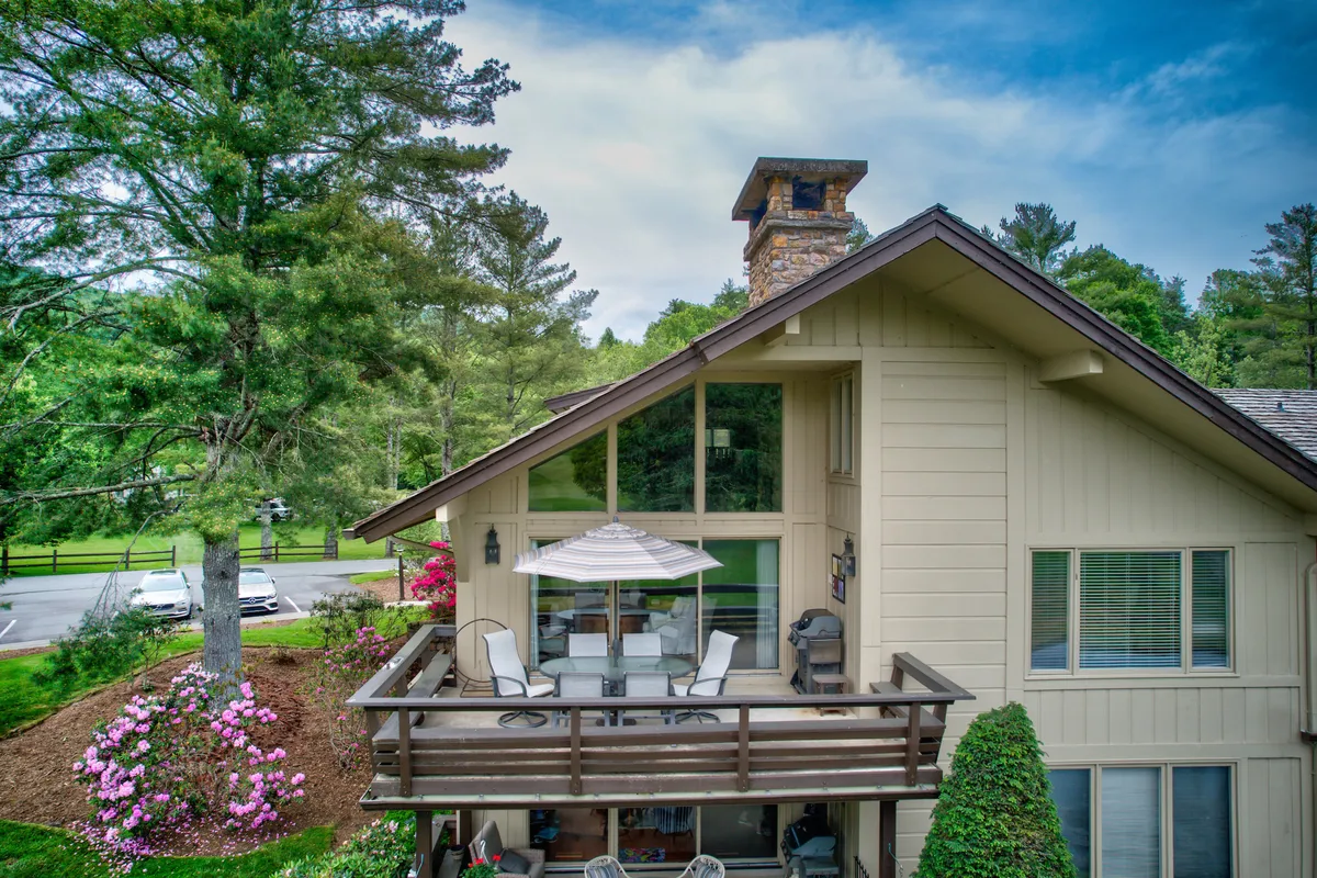 The image showcases a tranquil balcony with comfortable seating, a prominent chimney, and large windows framing views of the surrounding greenery. The space feels open and inviting, ideal for outdoor dining, relaxing, or enjoying the fresh mountain air as a group.
