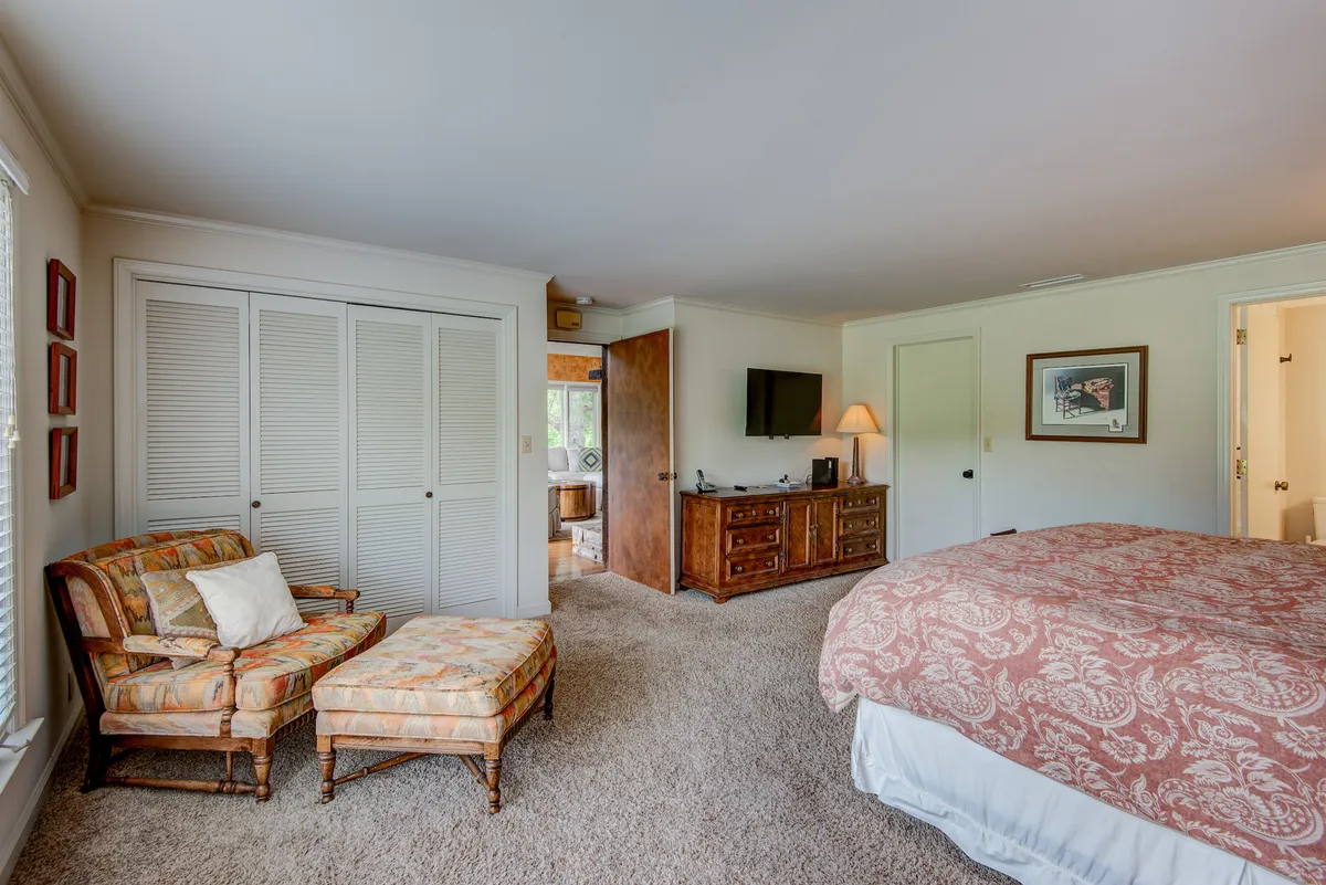 A cozy bedroom featuring a bed with patterned bedding, an inviting armchair and ottoman, a wooden dresser, and framed artwork on the wall. The setting is bathed in natural light, enhancing the sense of warmth and comfort.
