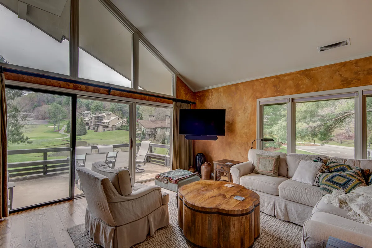 This image features a cozy living room with large glass windows, a wooden coffee table, plush seating, and decorative cushions. The room offers uninterrupted views of the outdoors and a relaxing, social ambiance.