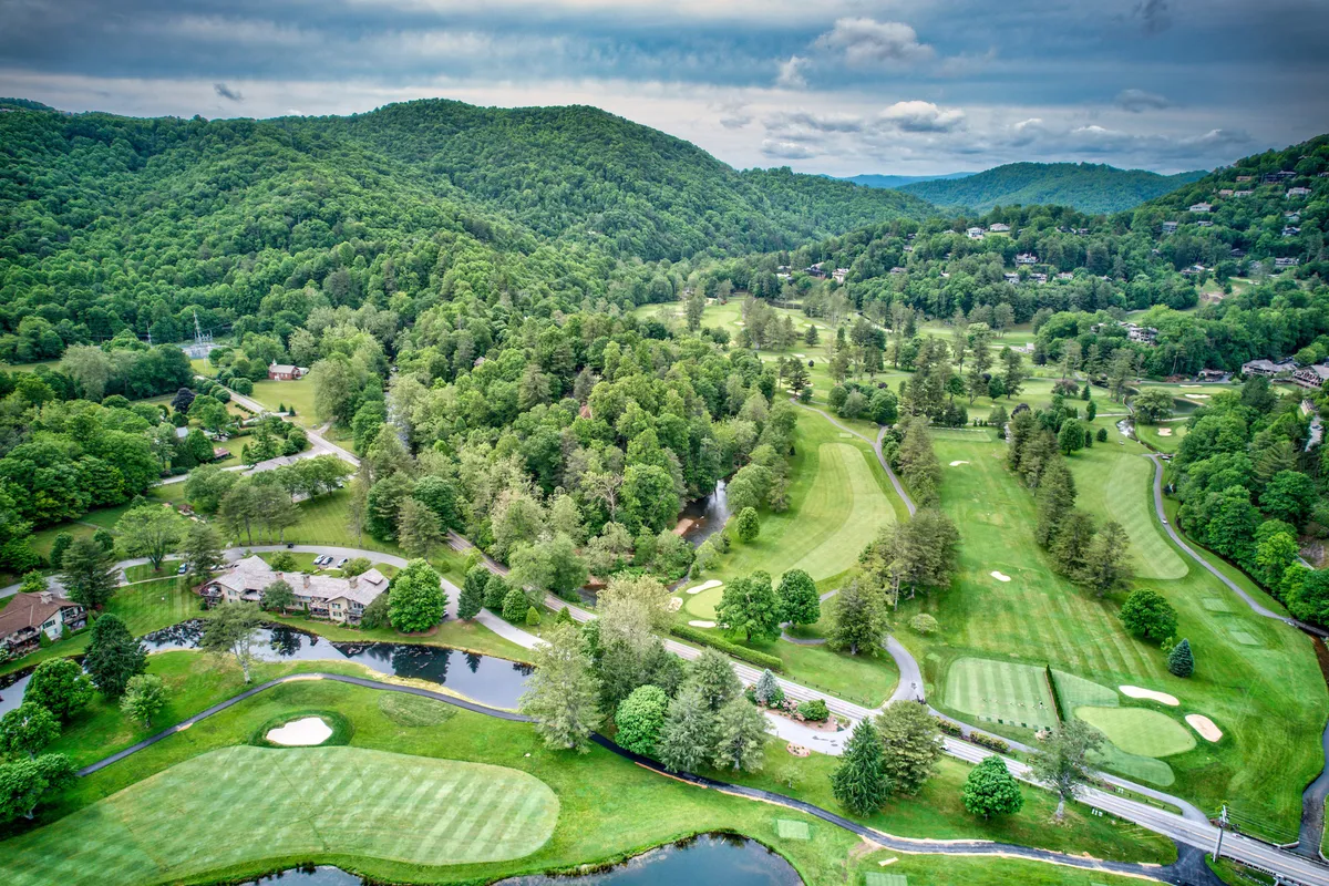 An expansive view of lush green mountains, a meandering river, a golf course, and scattered residential properties. The scene is framed by open sky, offering a panoramic glimpse of the natural beauty and recreational opportunities available within the community.