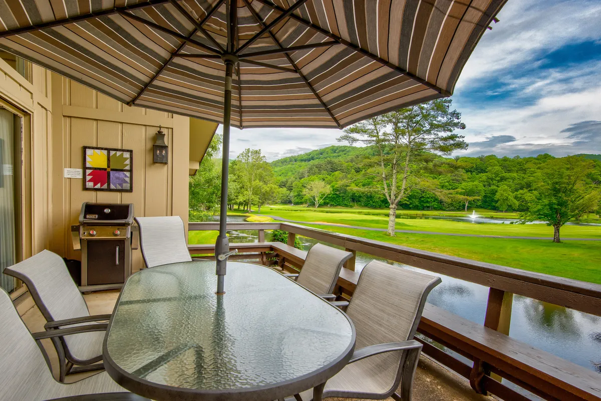 An outdoor dining area with a glass-top table, striped umbrella, and a grill overlooking a tranquil landscape. The setting invites relaxed socializing and al fresco dining with beautiful views.