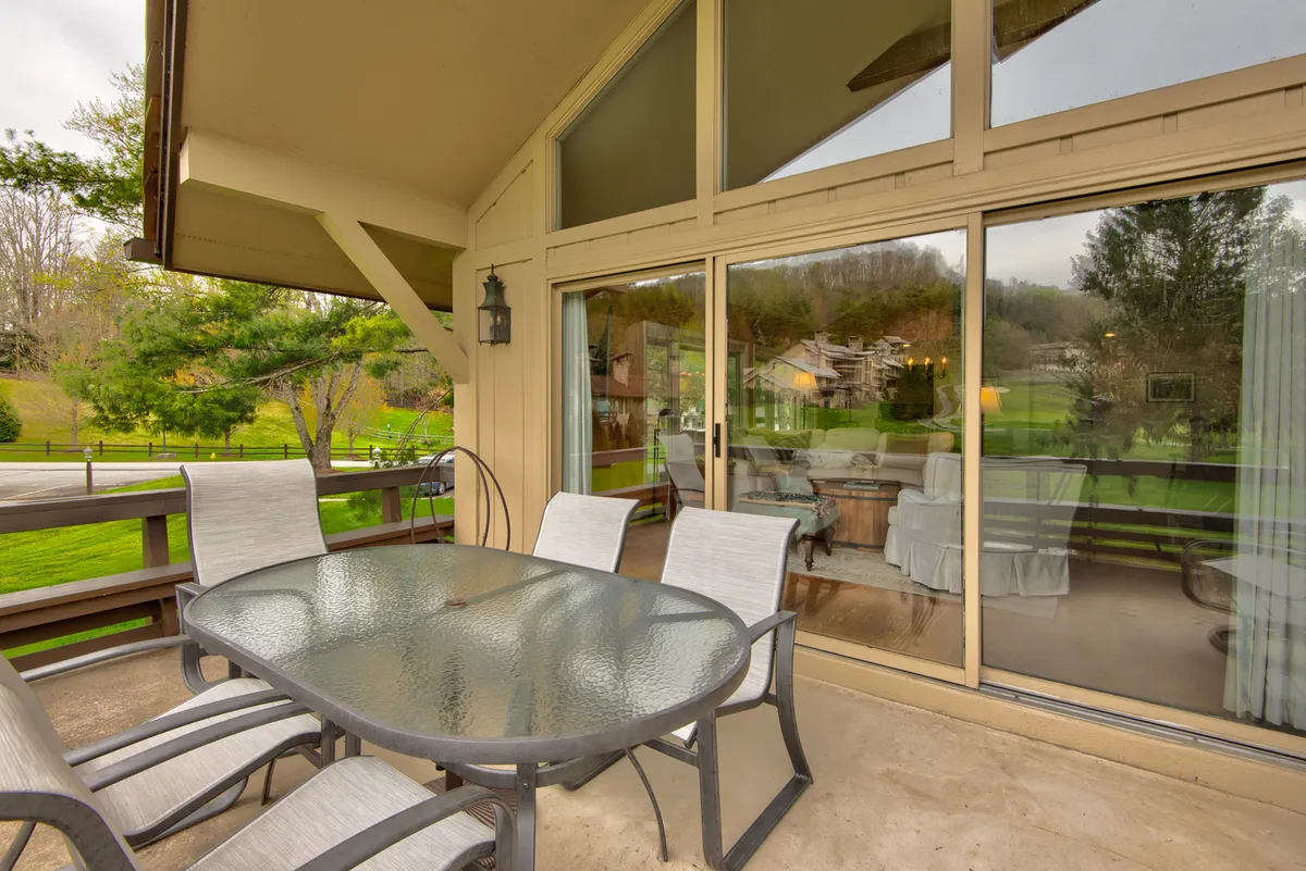 An exterior shot featuring an outdoor dining table on a deck beside large sliding glass doors, surrounded by vibrant greenery. The setting is inviting and highlights a comfortable space for gatherings, meals, and relaxation with beautiful views.