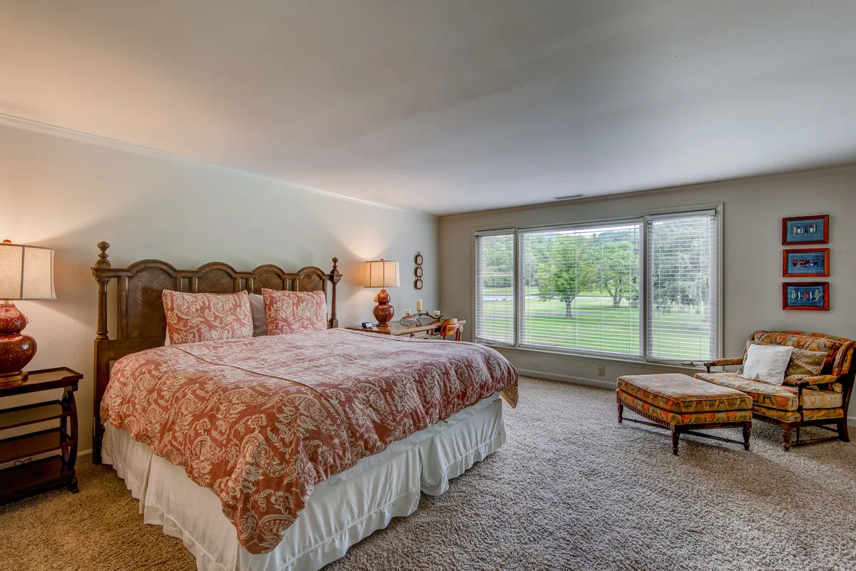 A spacious king bedroom with a large bed adorned in patterned bedding, expansive windows offering serene views of the outdoors, and a comfortable armchair positioned for relaxation or reading. The room is imbued with natural light, creating a calm and inviting atmosphere.