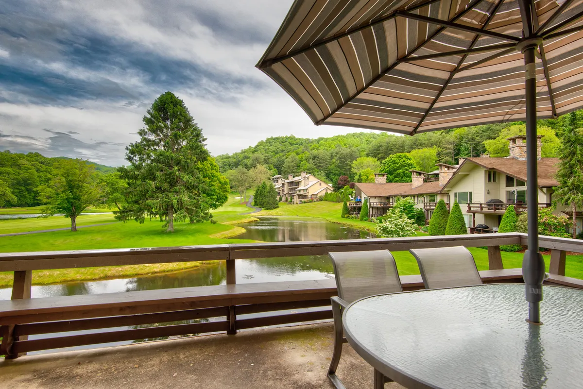 An exterior deck scene with a patio table and umbrella, overlooking the lake and surrounded by lush greenery. The view from the deck is tranquil, with the lake and rolling hills visible in the distance.